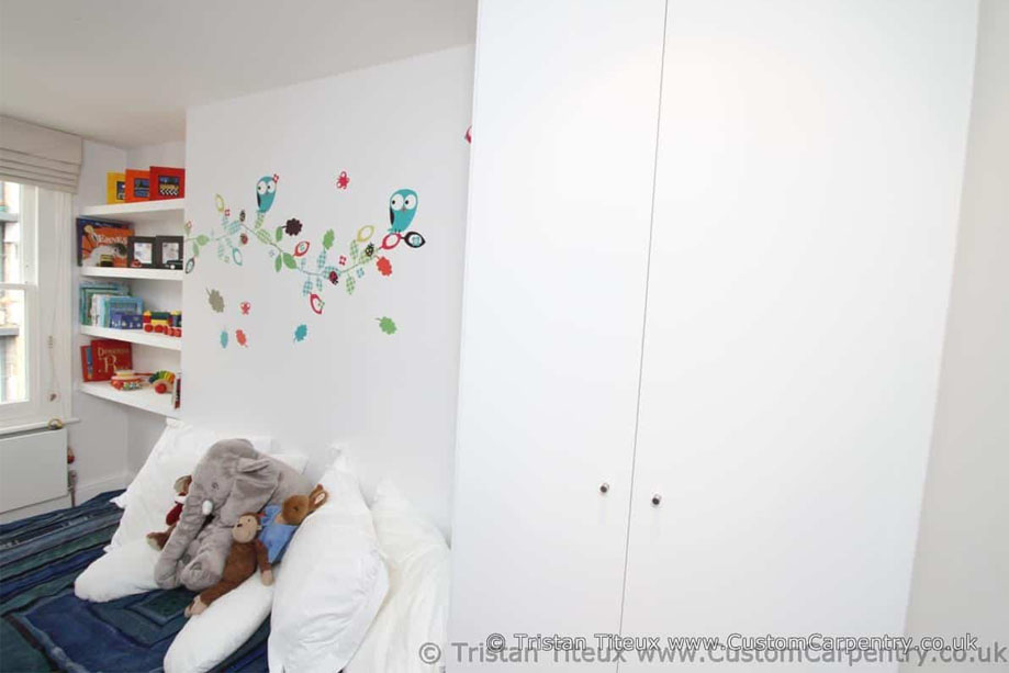 Interiors of children's room with wall recessed shelves by the window and tall fitted wardrobe next to a bed