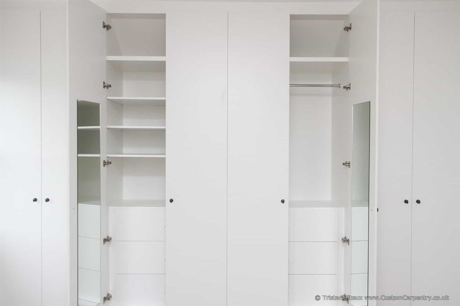 Open-door view of fitted floor to ceiling wardrobes painted White with custom shelving, drawers, clothes rack, and mirrors inside